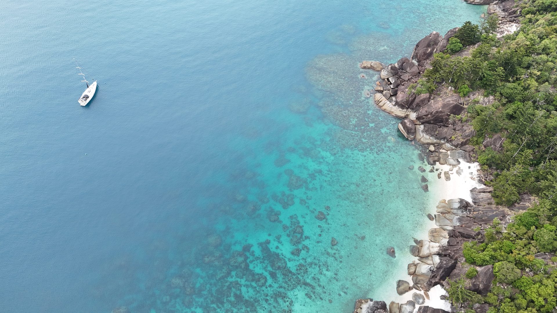 Shimbiri anchored alone off a turquoise reef-fringed shore