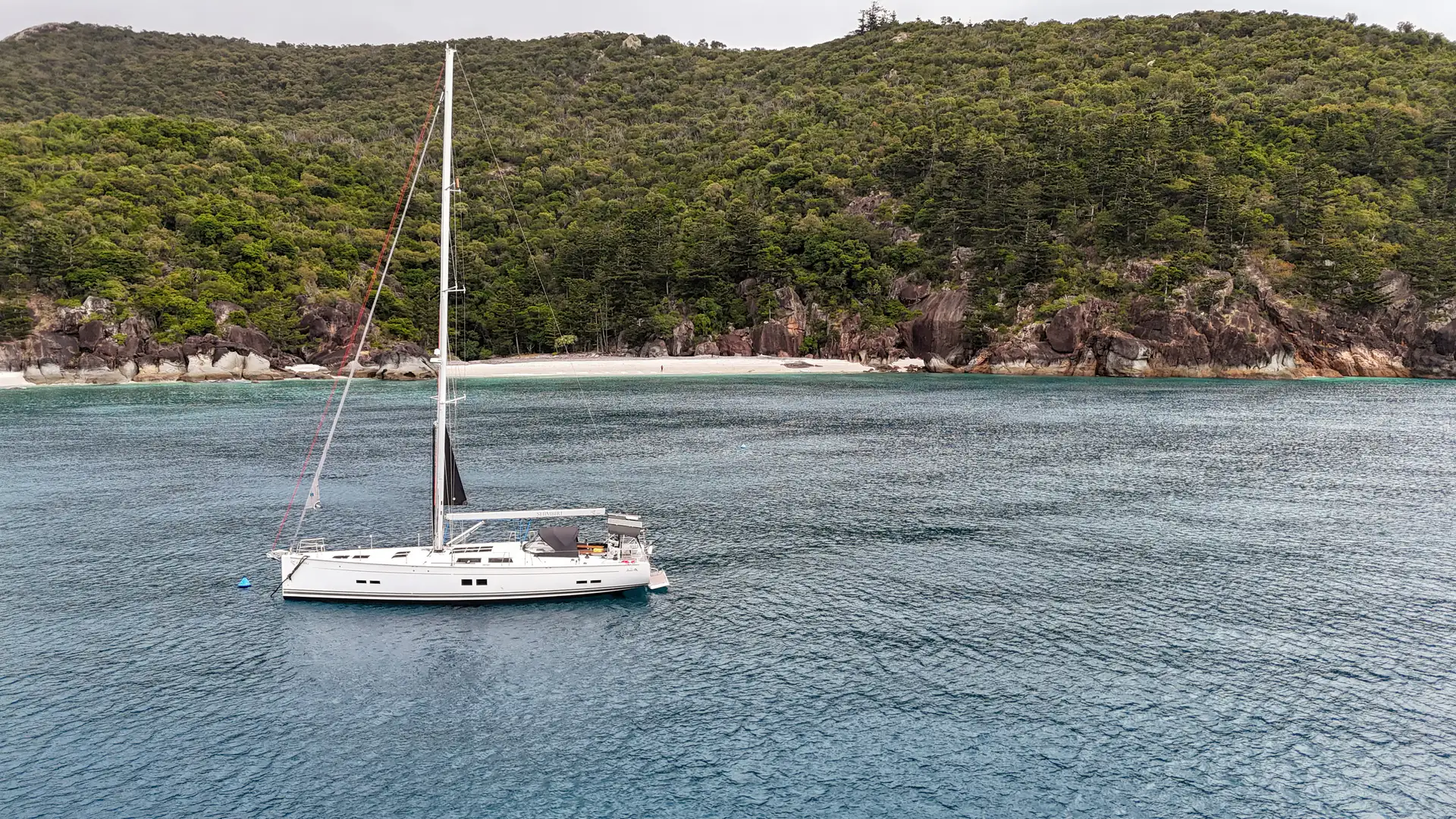 Shimbiri at anchor in a turquoise bay below a forested headland